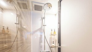 Moderni kylpyhuone, jossa on lasinen suihkuseinä, käsisuihku ja seinään asennettu hana. Hyllyllä on kaksi shampoopulloa.