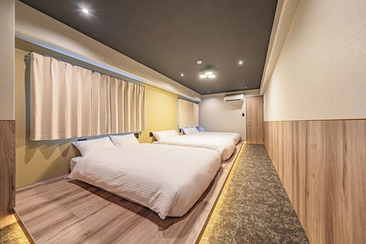 A hotel room with two beds, a wooden floor, a carpeted area, and an air conditioning unit.