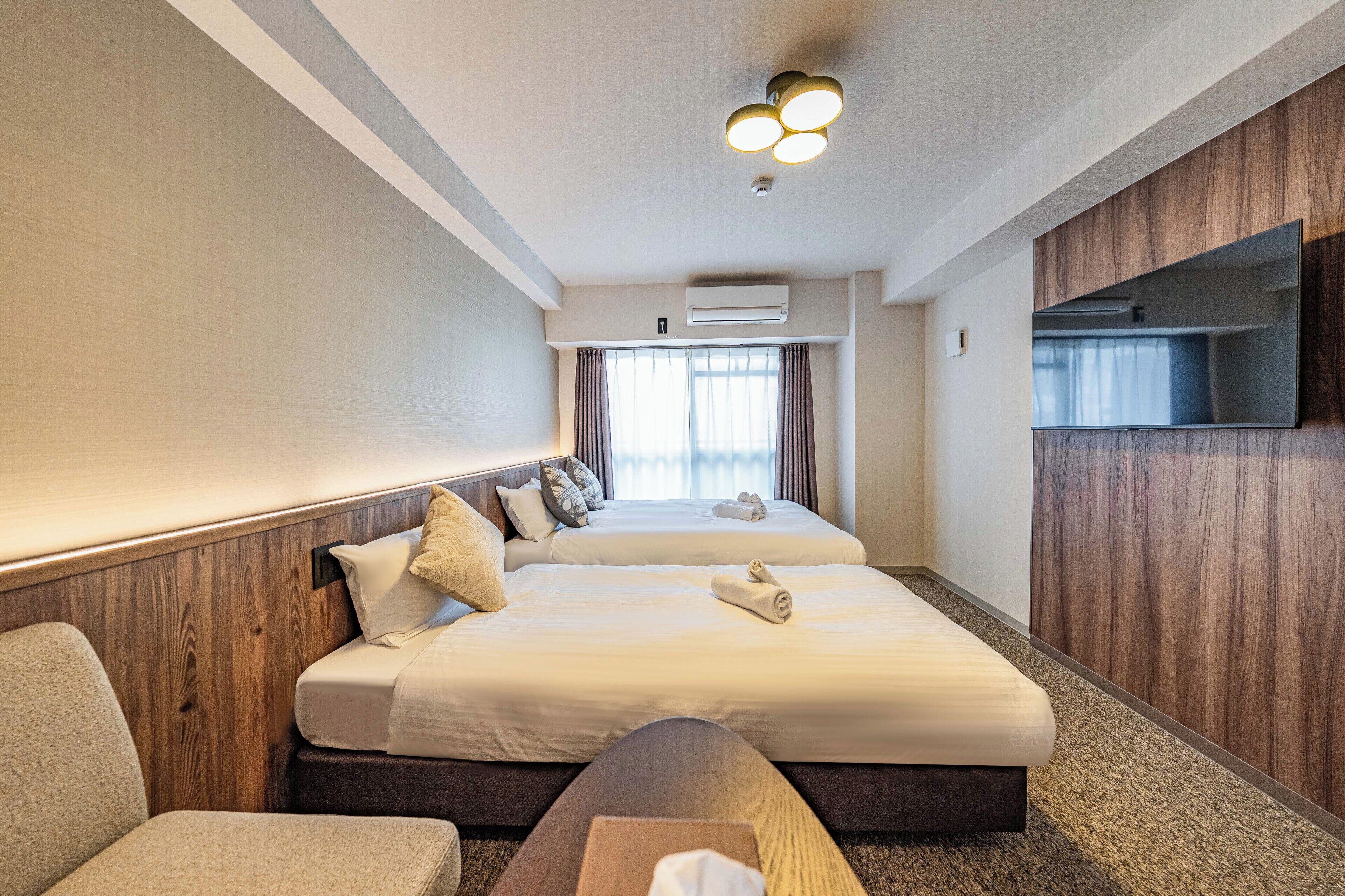 A hotel room with two beds, a sofa, a TV, and an air conditioning unit.