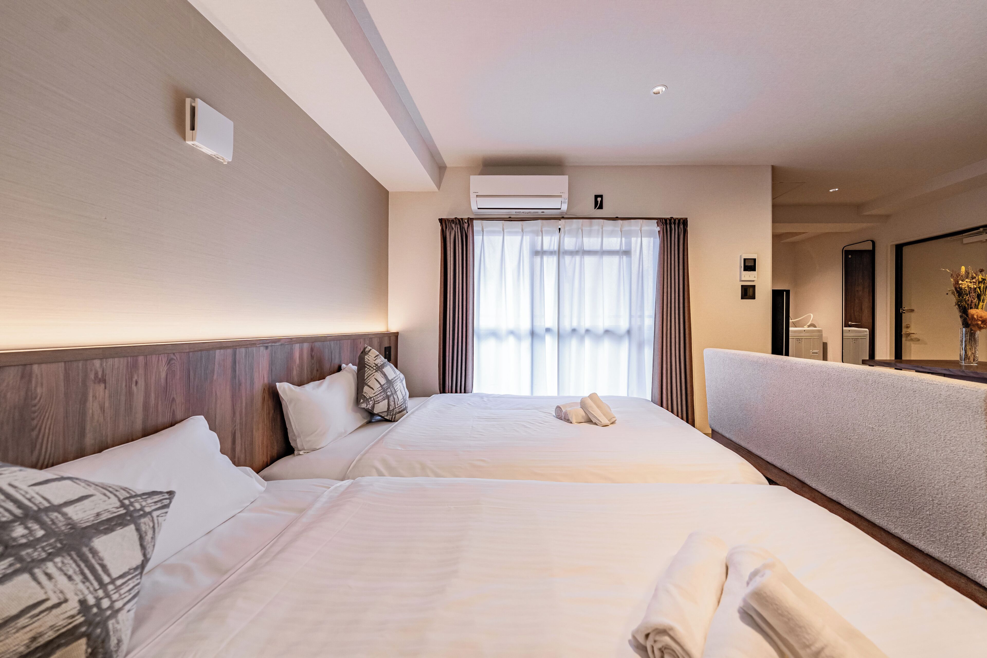 A hotel room with two beds, a large window with curtains, a wall-mounted air conditioner, and a mirror.