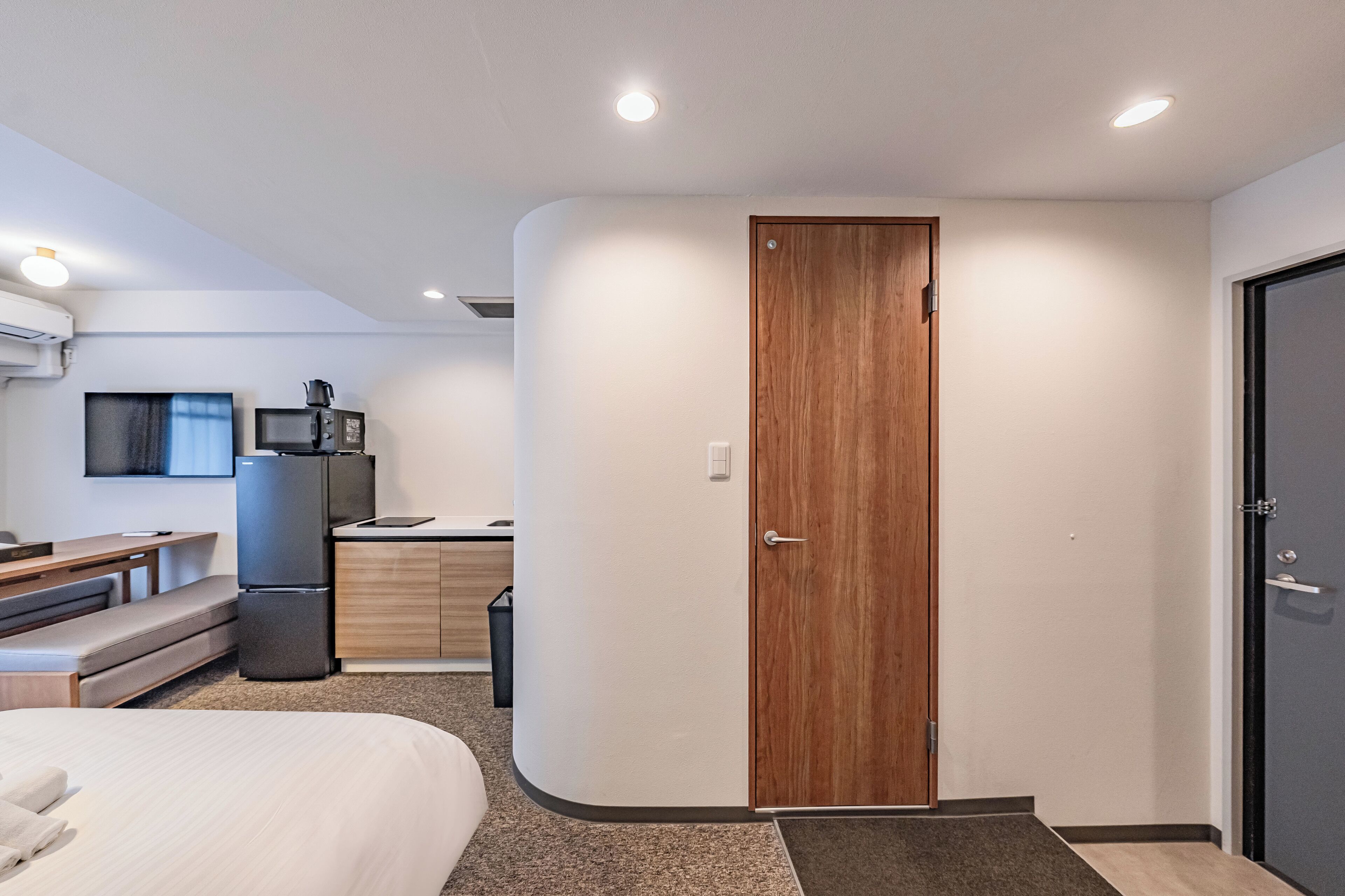 Een moderne hotelkamer met een bed, een bureau, een kitchenette en een houten deur.
