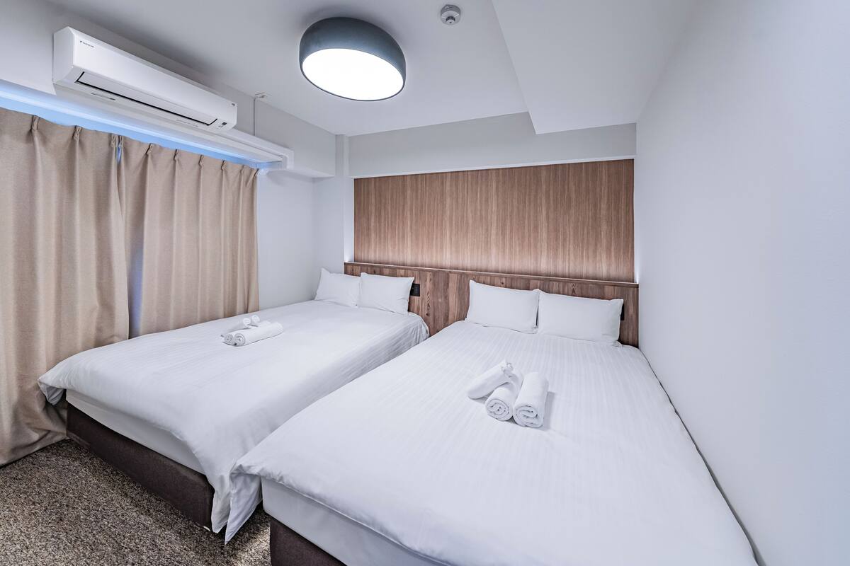 A hotel room with two beds, beige curtains, a wooden headboard, and an air conditioning unit.