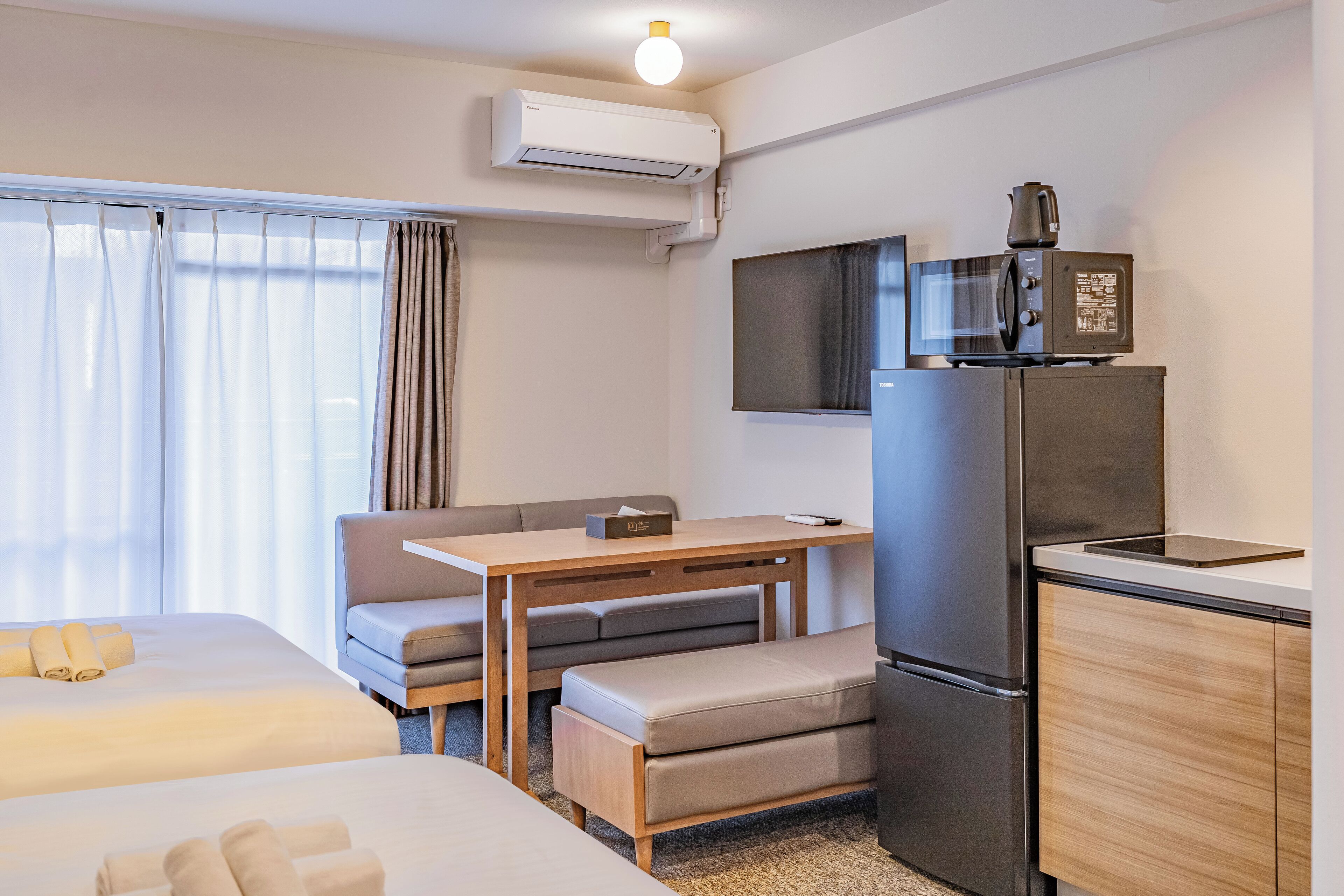 A compact hotel room with a bed, a desk, a chair, a sofa, a kitchenette, and an air conditioning unit.