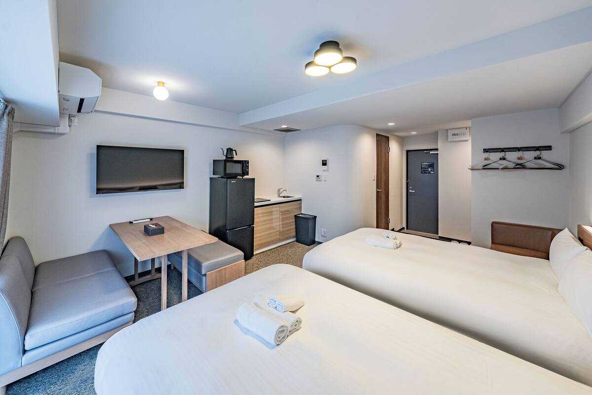 A hotel room with two beds, a desk, a TV, a microwave, and a small kitchenette.