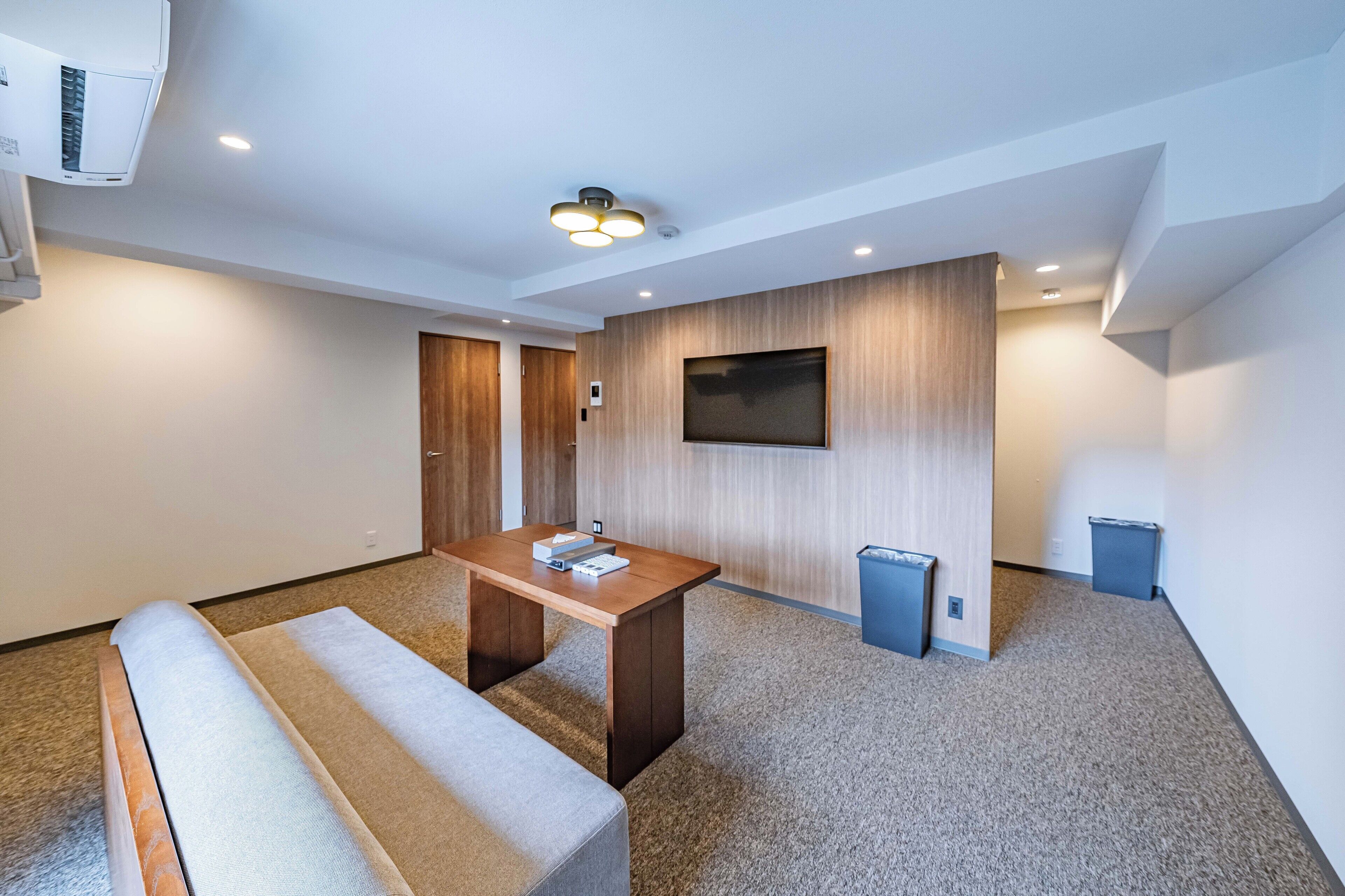 Een moderne hotelkamer met een houten bureau, een bankje, een flatscreen televisie aan de wand en een luchtverfrisser in het plafond.