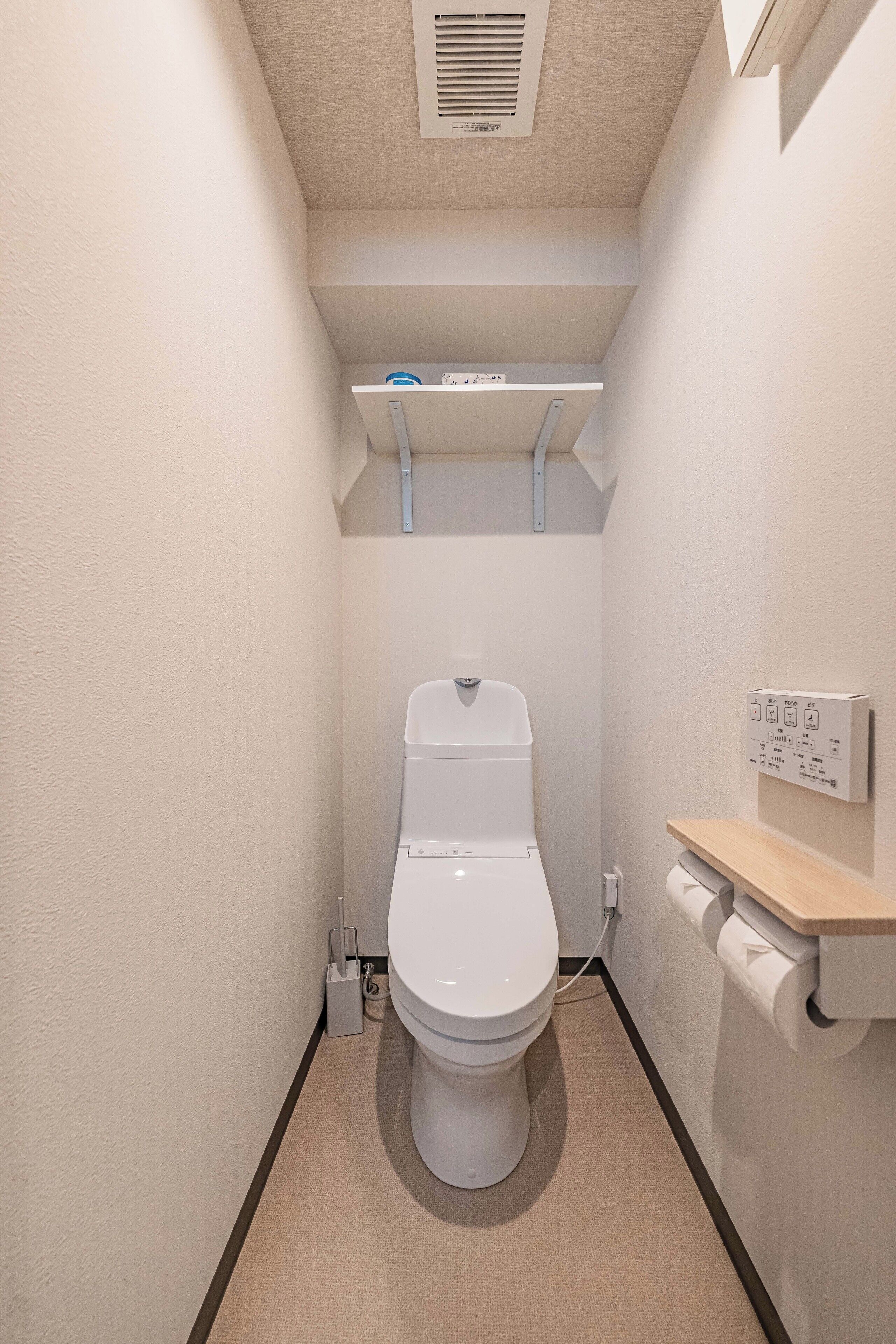 A compact bathroom with a white toilet, a small shelf above it, and a wall-mounted flush handle.