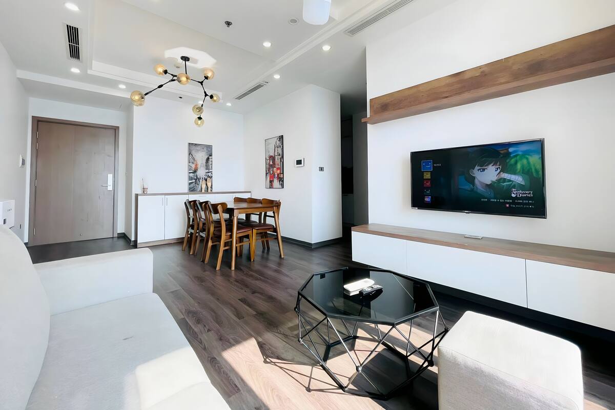 A modern living room with a glass coffee table, wooden dining table, and a TV mounted on the wall.