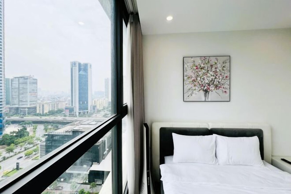 A hotel room with a bed, a window with a city view, and a framed artwork on the wall.