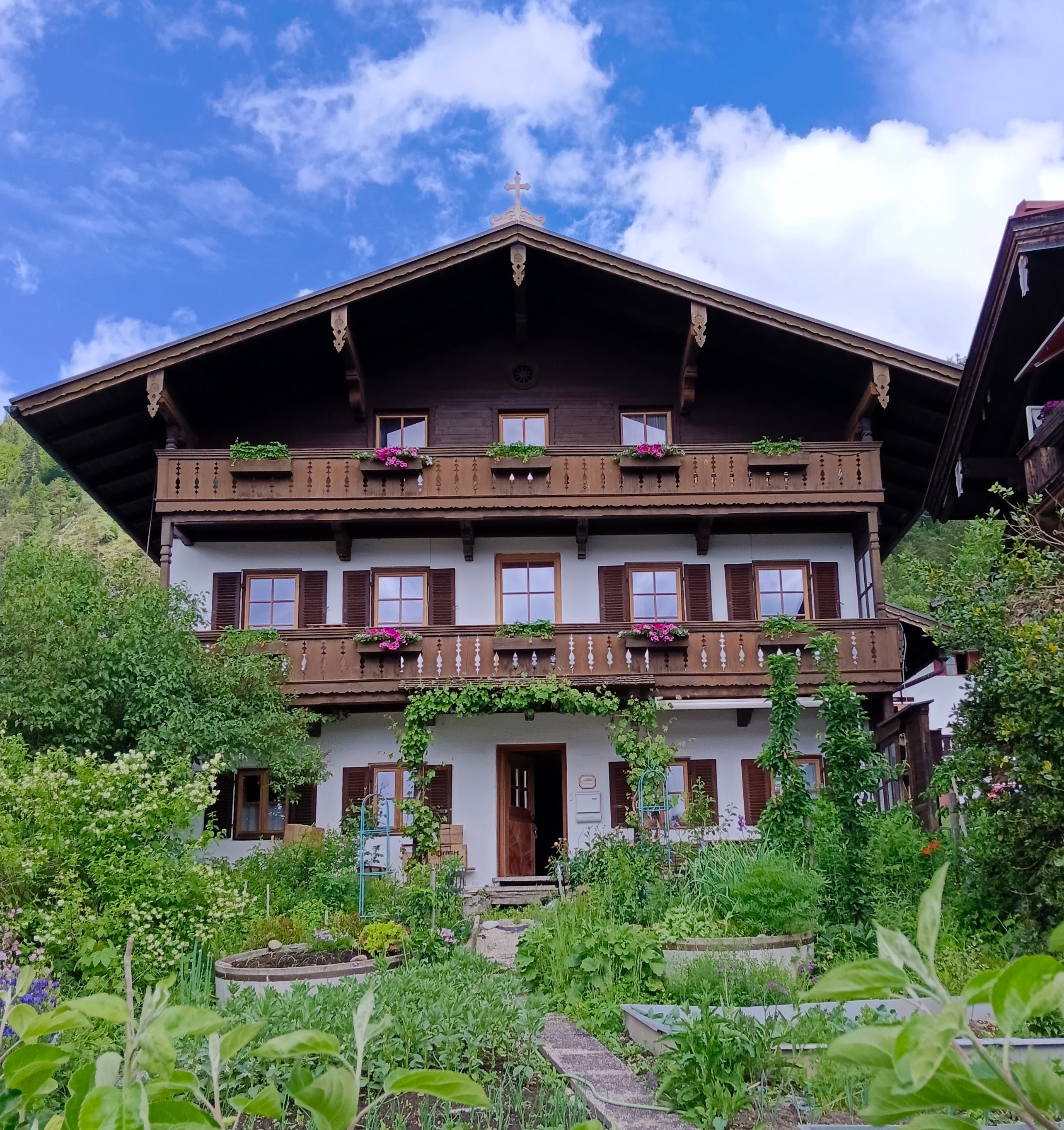 A two-story chalet with a balcony, surrounded by lush greenery and a clear sky.