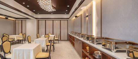 A hotel dining area with tables and chairs, a buffet line, and a decorative ceiling light fixture.