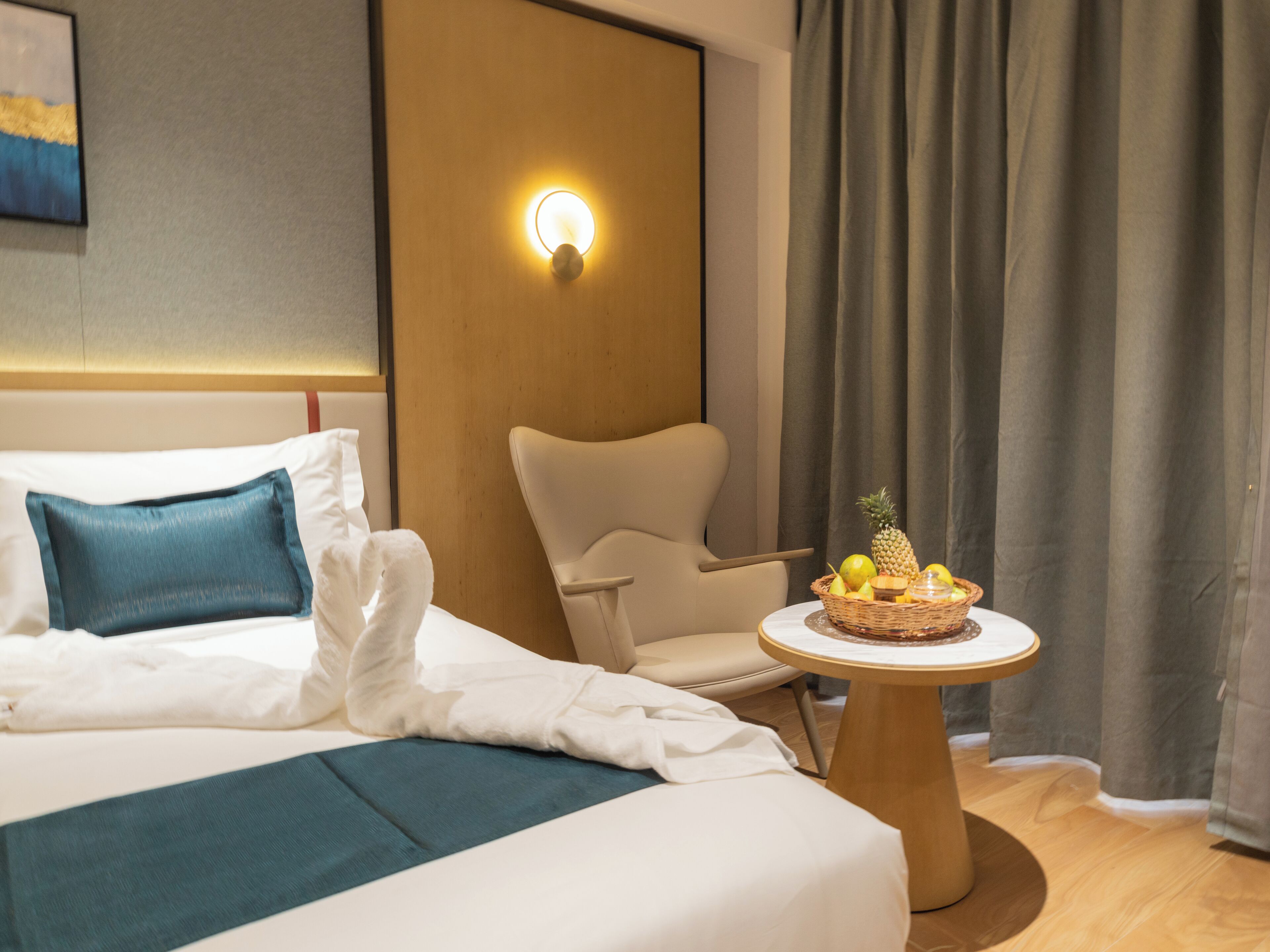 A hotel room with a bed, a chair, a table with a fruit basket, and a wall-mounted light.