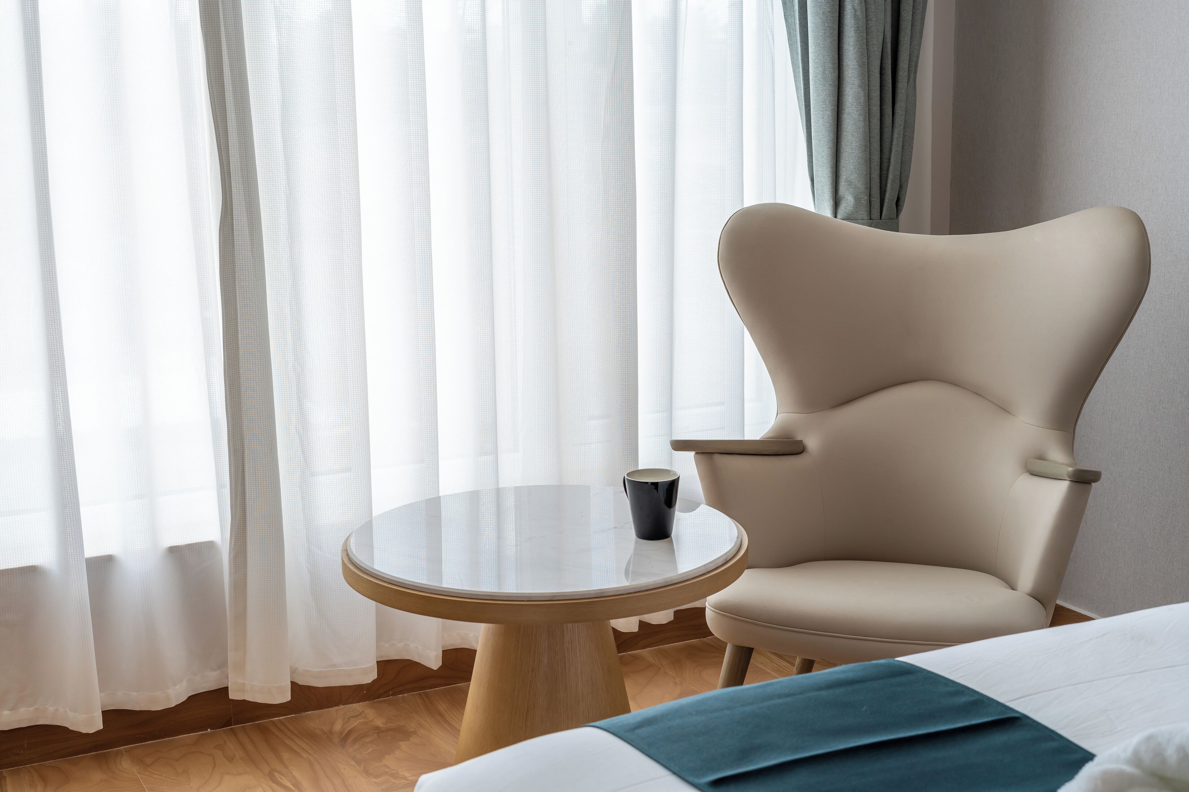 Ein Hotelzimmer mit einem runden Glastisch, einem beigen Sessel, einem Bett mit blauer Tagesdecke und transparenten Vorhängen.