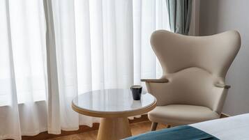 Ein Hotelzimmer mit einem runden Glastisch, einem beigen Sessel, einem Bett mit blauer Tagesdecke und transparenten Vorhängen.