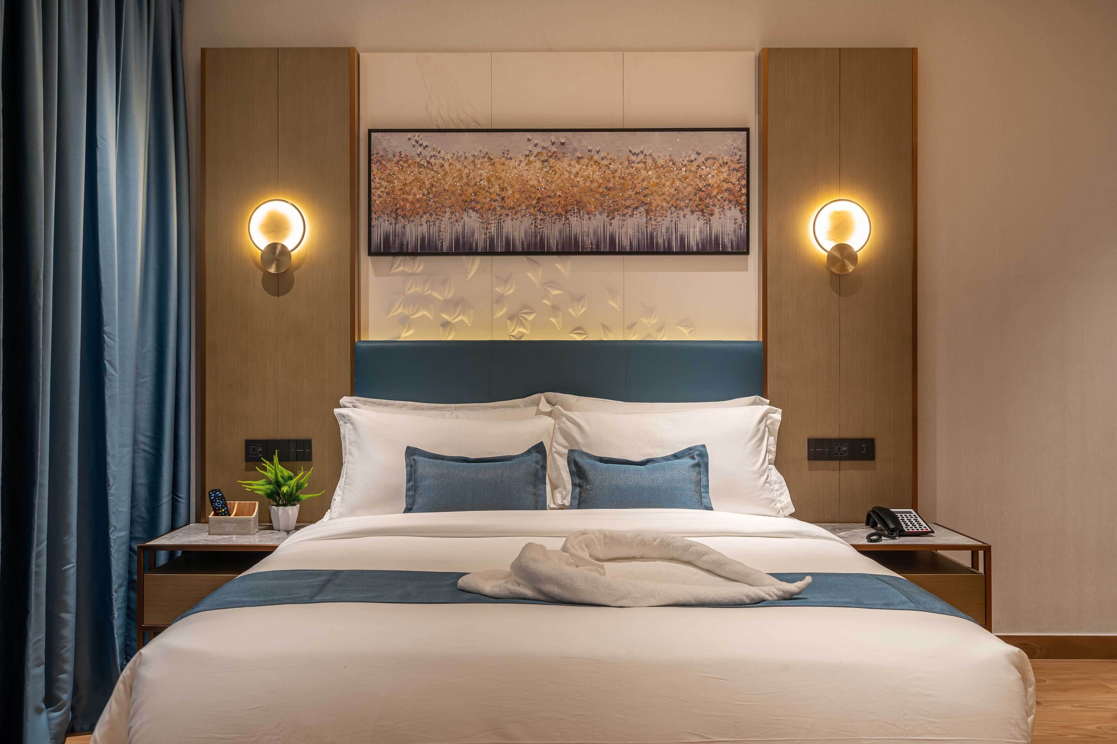 Una cama bien hecha con edredón blanco y almohadas azules, una toalla blanca sobre la cama y un cuadro en la pared superior. Hay dos mesitas de noche, una de ellas con una lámpara y un teléfono.