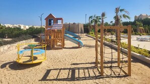 A sandy playground with a slide, climbing structures, and a small house-like playhouse.