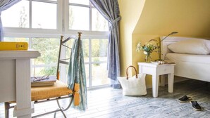 Une pièce avec une grande fenêtre, un lit blanc, une petite table de chevet ornée d’un vase de fleurs, une chaise avec un coussin et une paire de chaussures posées sur le sol.
