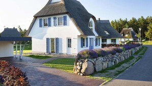 Une maison blanche avec un toit de chaume, un mur de pierres orné de fleurs et une allée pavée.