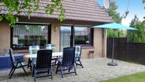 A patio with a table and chairs, a blue umbrella, and a brick house with a tiled roof. - Ferienhaus mit Garten in Nessmersiel (Neßmersiel)