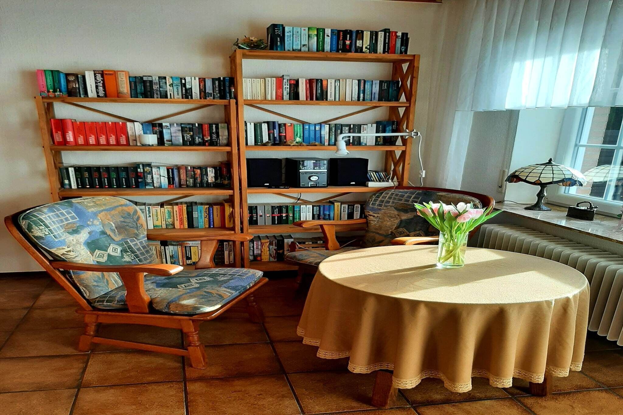 Ein Zimmer mit rundem Tisch, zwei Stühlen, einem Bücherregal und einem Heizkörper.
