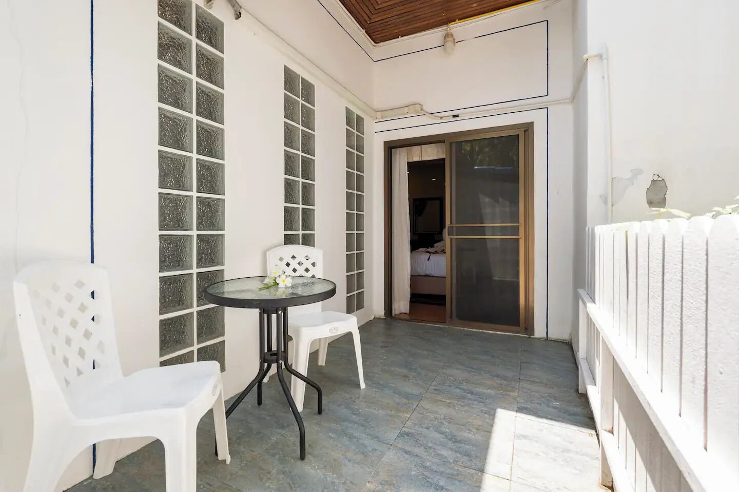 A small patio with a round table and two white chairs, a sliding glass door leading to a bedroom, and a white fence along the right side.