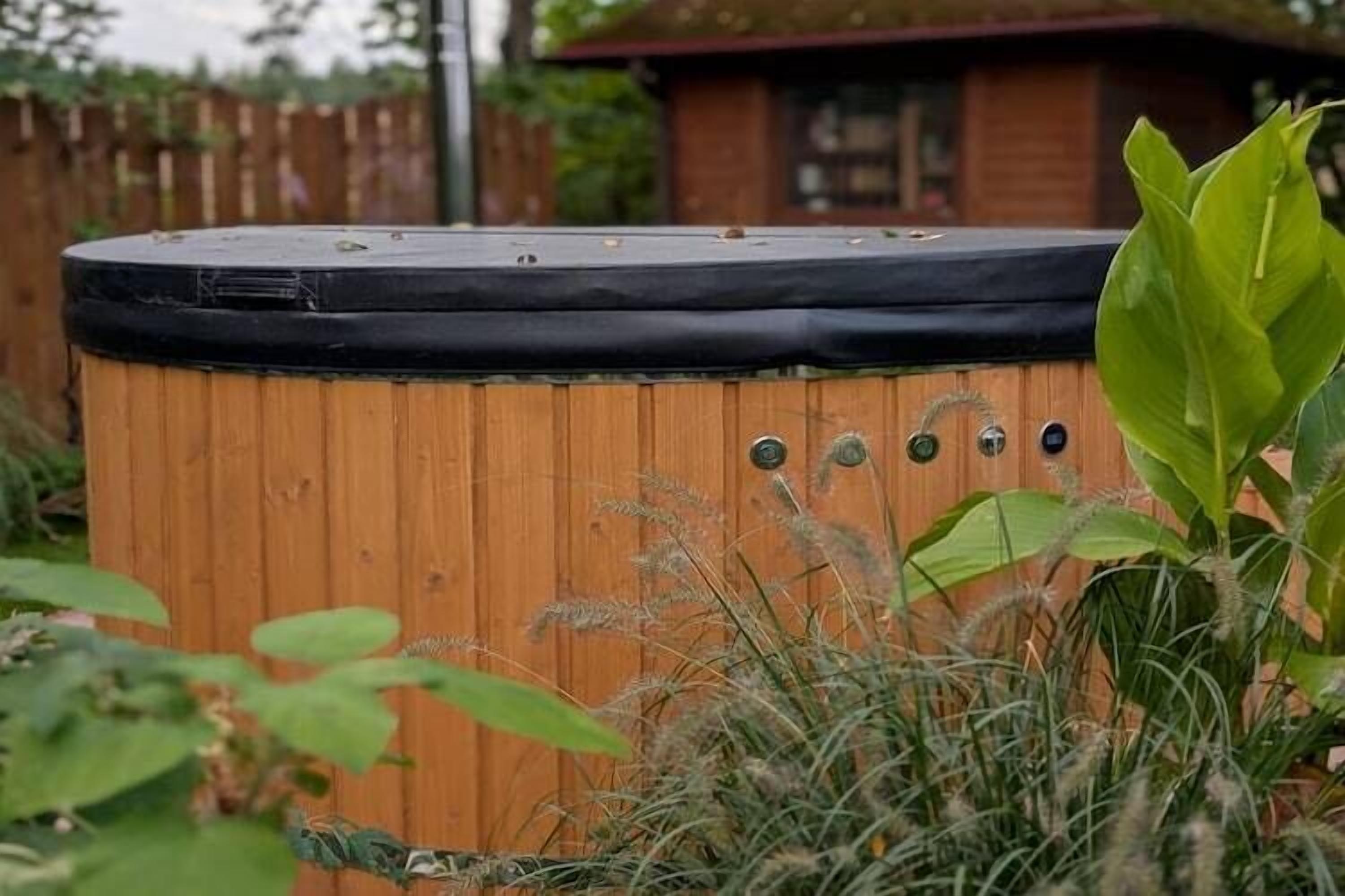 Un bain à remous en bois, surmonté d’un couvercle noir, entouré de verdure et bordé d’une clôture en bois à l’arrière-plan.