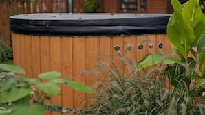 A wooden hot tub with a black cover, surrounded by greenery and a wooden fence in the background. - Holiday home with jacuzzi and private garden (Gizycko)