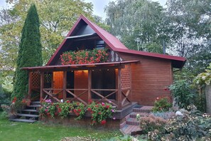 A wooden cabin with a sloped roof, a porch with lattice railings, and a garden with flowering plants. - Holiday home with jacuzzi and private garden (Gizycko)