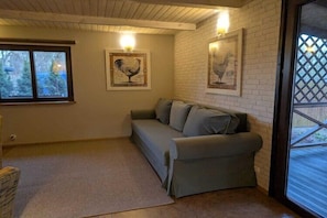 Une pièce avec un canapé gris, un fauteuil en osier, une fenêtre avec des stores et deux appliques murales.