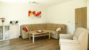 A living room with a beige sofa, a wooden coffee table, and a cream-colored armchair. There is a cabinet with glass doors on the left and a door on the right.