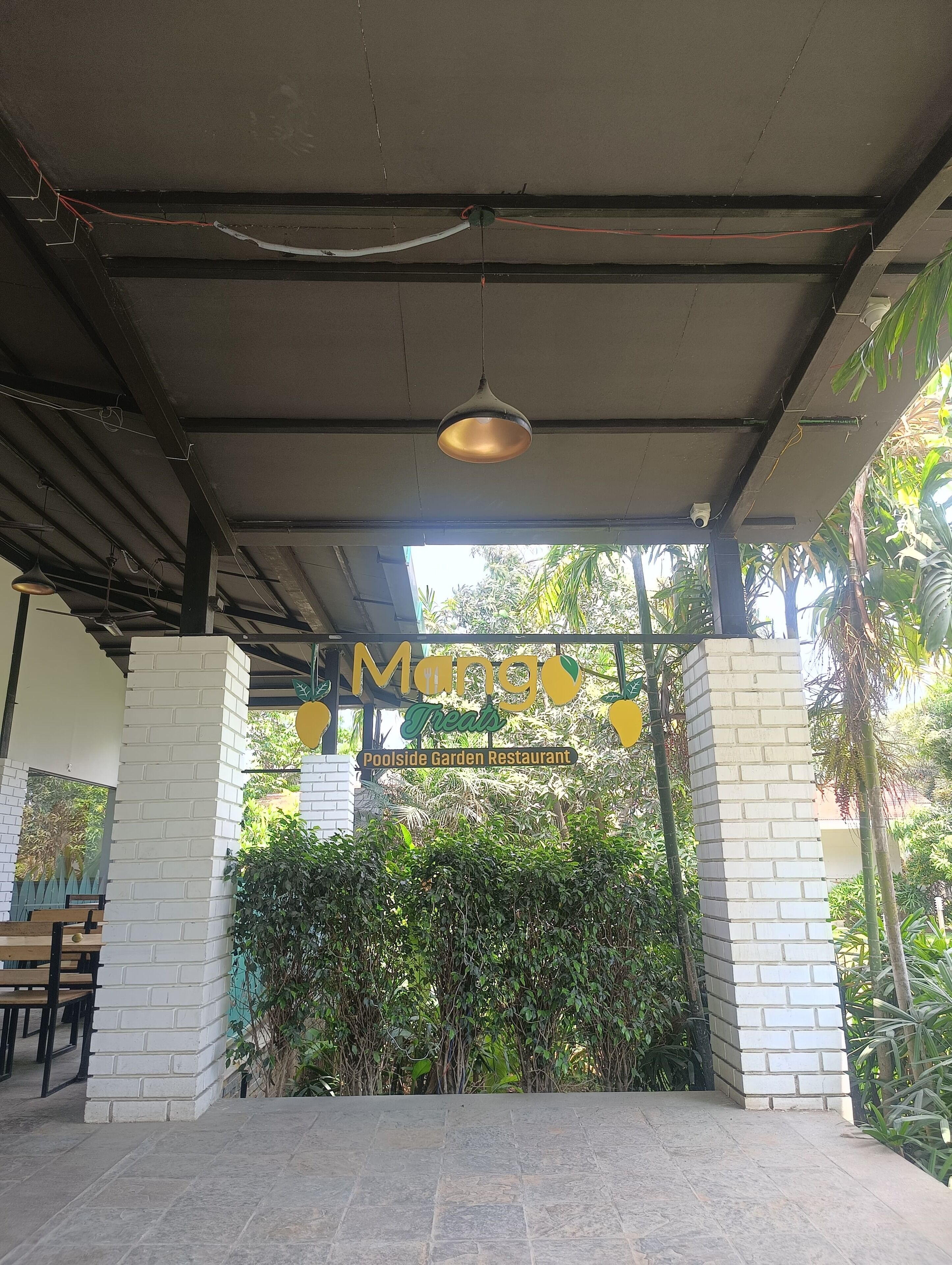 Un espace extérieur abrité, agrémenté d’une enseigne indiquant « Mango Treats Poolside Garden Restaurant », entouré de verdure et délimité par un mur de briques.