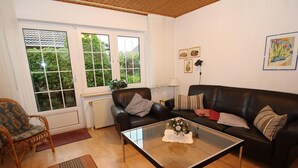 Een woonkamer met een glazen salontafel, twee zwarte leren banken, een rieten fauteuil en uitzicht op groen via grote ramen.