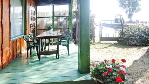 Un pontile in legno con tavolo e sedie, una pianta in vaso con fiori rossi e bianchi, e vista su un corpo d'acqua e alberi.