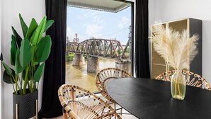 Ein Zimmer mit einem großen Fenster mit Blick auf eine Brücke, einem schwarzen Tisch, Korbstühlen, einer Topfpflanze und einer Vase mit Pampasgras.
