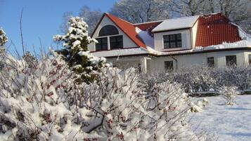 Une maison avec un toit rouge et des murs blancs, entourée d’arbustes et d’arbres couverts de neige.