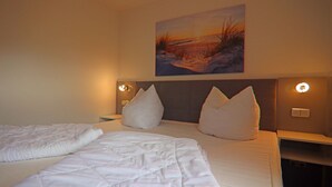 A hotel room with a large bed, two pillows, a nightstand, a wall-mounted painting, and a wall socket. - Appartementhaus Nicole, Zinnowitz (Zinnowitz)