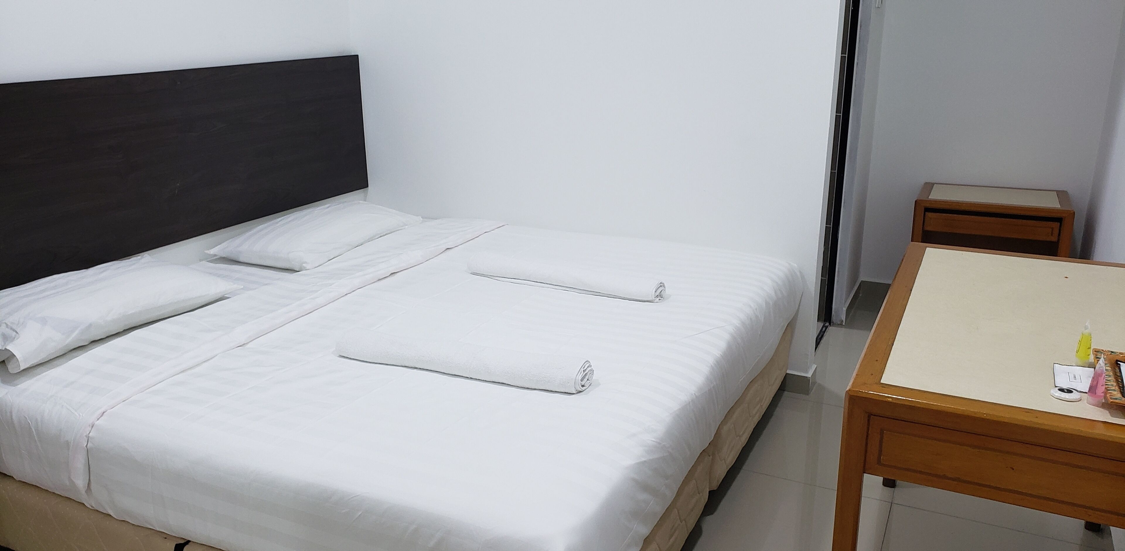A neatly made bed with white linens and two rolled towels, a wooden nightstand with toiletries, and a small table with a white top.