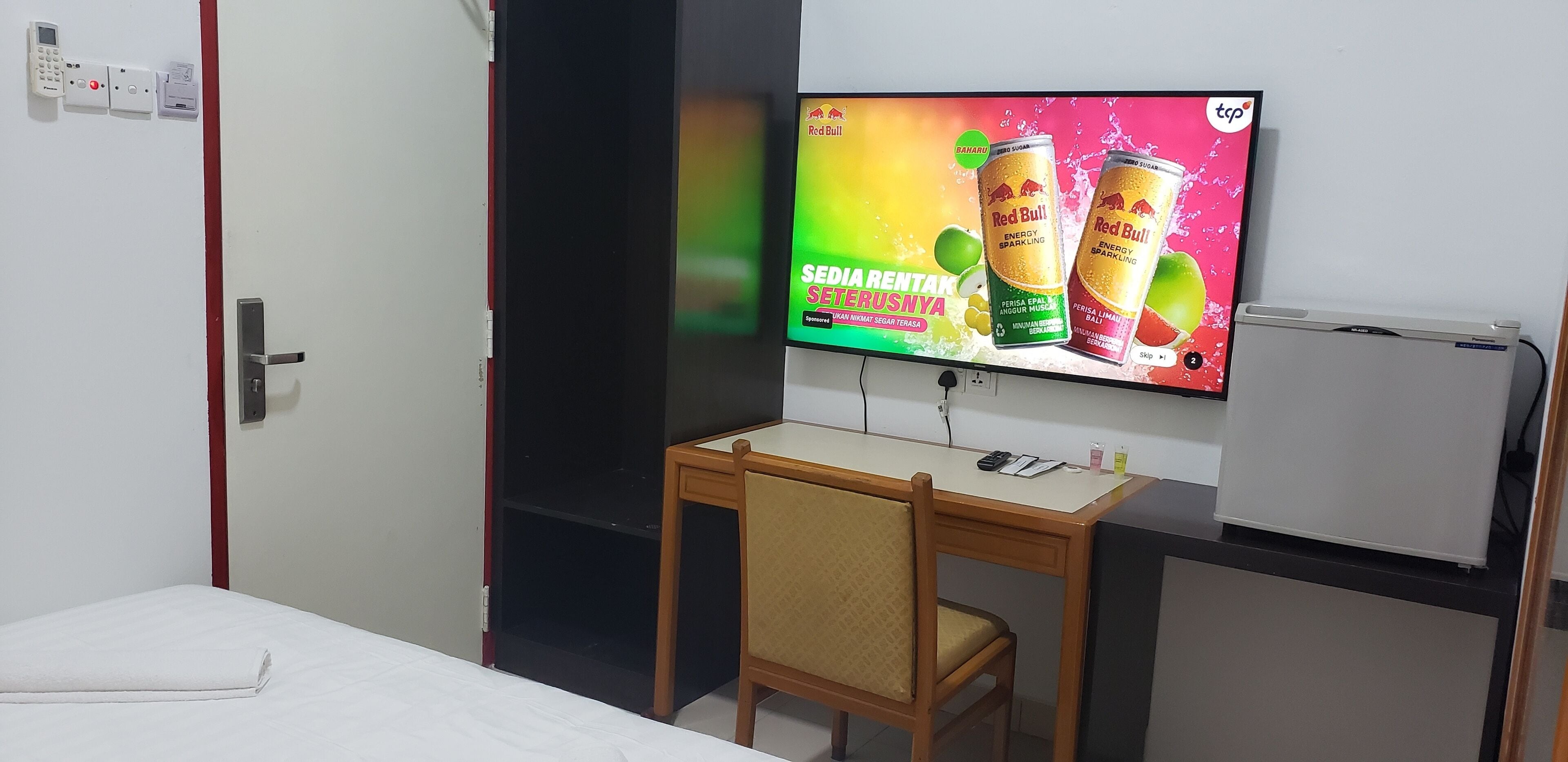 A hotel room with a bed, a desk, a chair, a television displaying an advertisement, and a whiteboard.