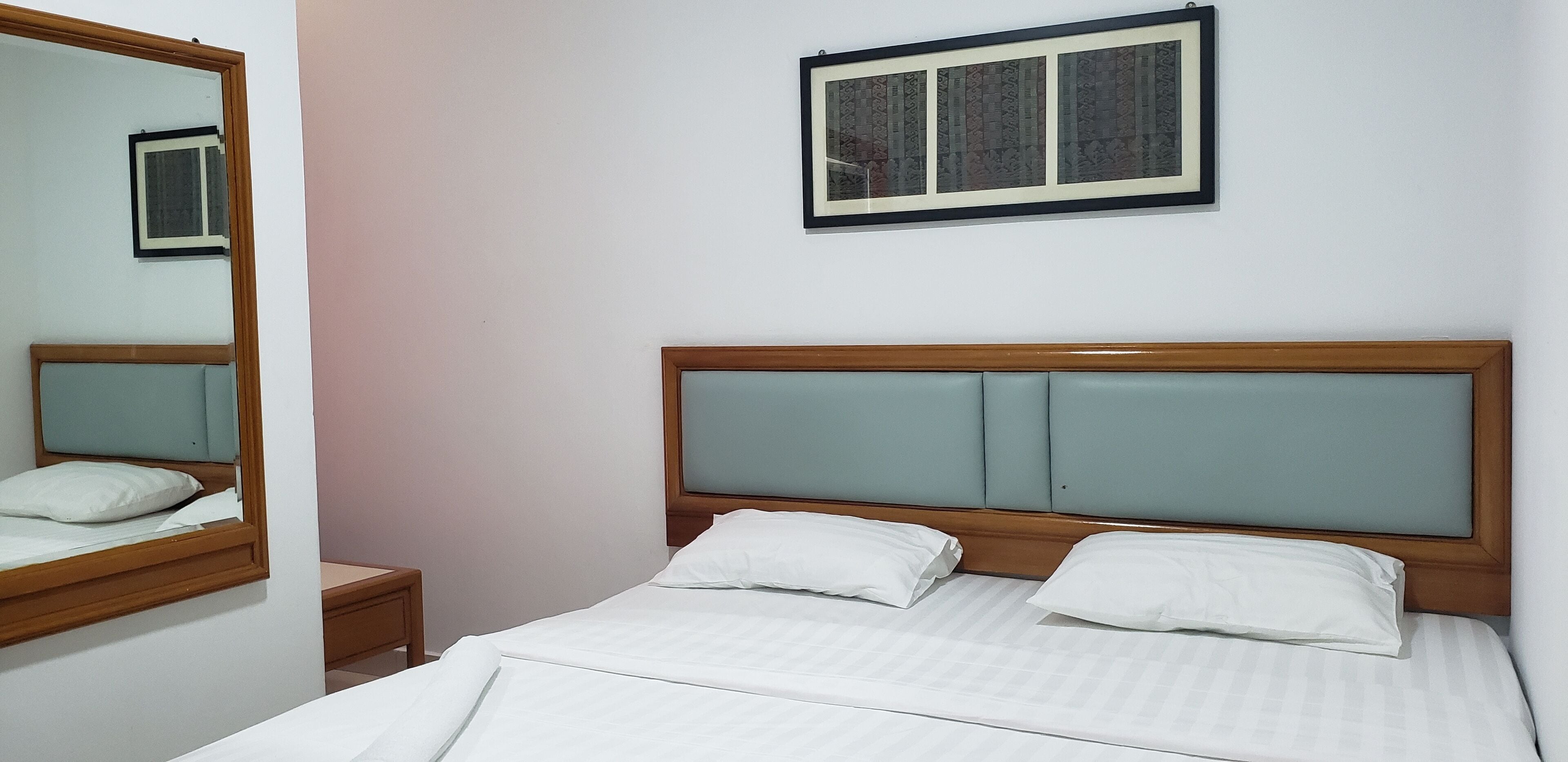 Un letto rifatto con cura, con una testiera in legno, due cuscini, un comodino e uno specchio appeso al muro.