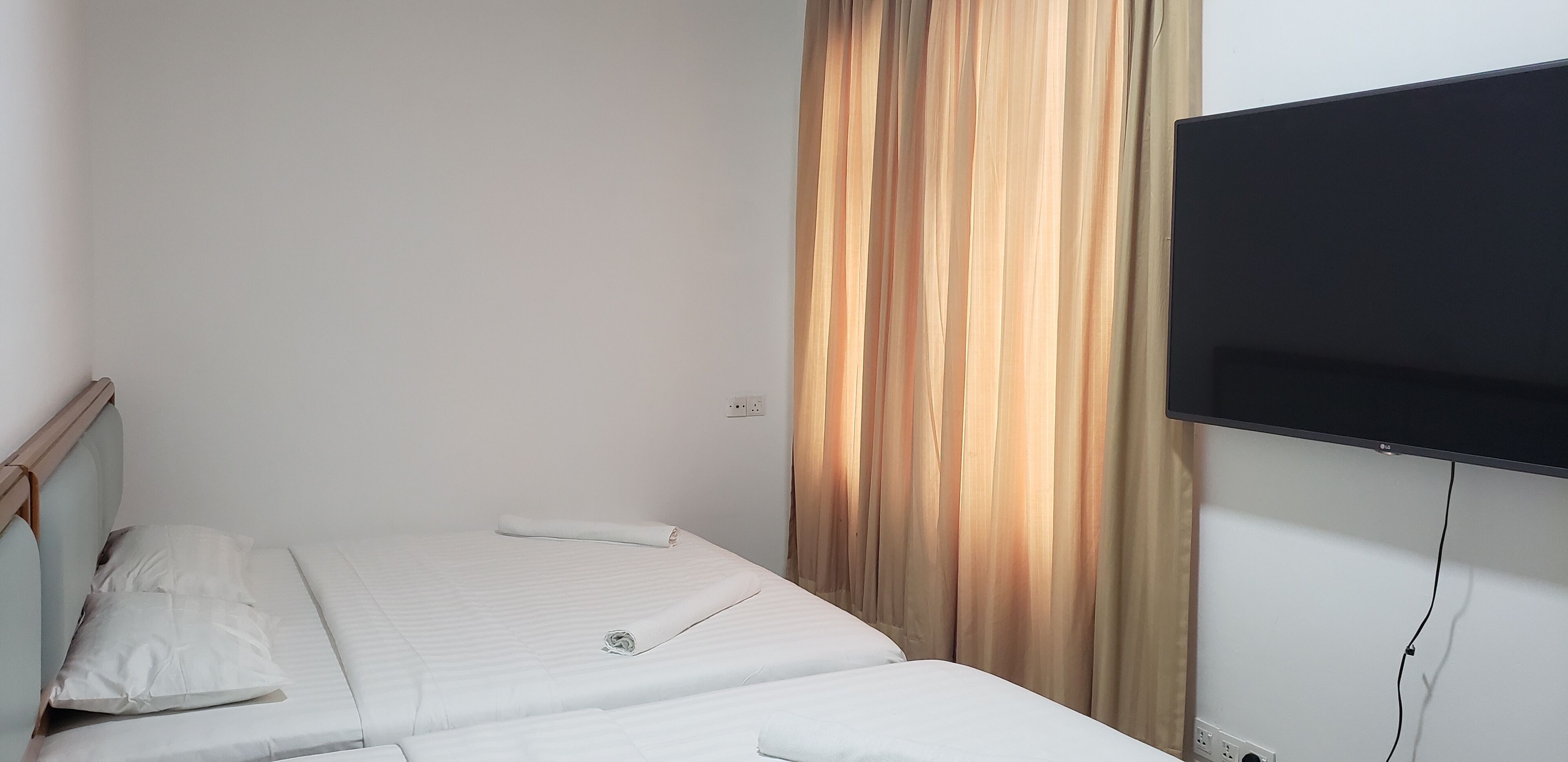 Una cama individual con ropa de cama blanca y cabecera de madera, un televisor montado en la pared y una cortina con estampado de rayas verticales.