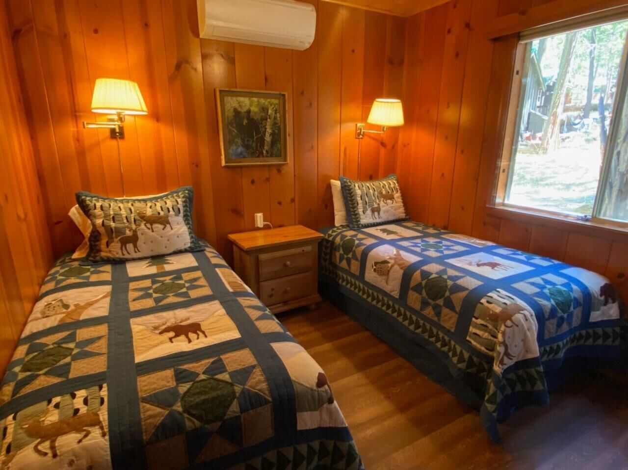 A cabin with two wooden beds, each with a quilted blanket and a matching pillow. There is a nightstand between the beds, and a window on the right side.