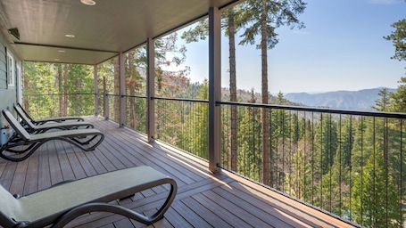 A wooden deck with two lounge chairs and a covered area, overlooking a forest and mountains.