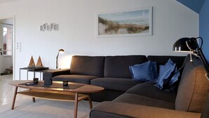 A modern living room with a sectional sofa, a coffee table, and a side table with a lamp. There is a framed picture on the wall and a small shelf with decorative items. - Ferienhaus mit drei Bädern in Dornumersiel (Dornumersiel)