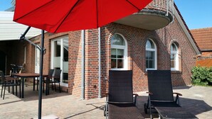 A patio area with two wicker chairs, a red umbrella, and a brick building with arched windows. - Ferienhaus mit drei Bädern in Dornumersiel (Dornumersiel)
