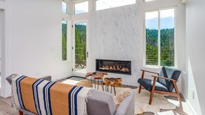 Ein geräumiges Wohnzimmer mit Kamin, großen Fenstern und Blick auf einen Wald.