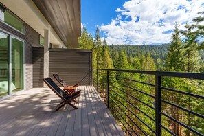 A wooden deck with two lounge chairs, a metal railing, and a view of a forested mountain range.