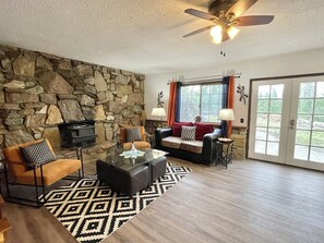 A living room with a stone wall, a fireplace, a sofa, a coffee table, and a ceiling fan. - Casa De Colores (Oakhurst)