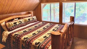 Een slaapkamer met een schuine kap, houten wanden, een bed met beddengoed met een patroon, een plafondventilator en ramen met uitzicht op een bos.