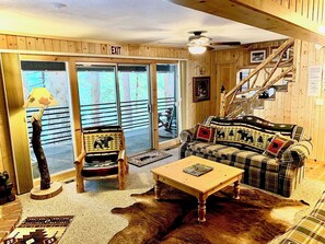 A cozy cabin interior with a plaid sofa, a wooden coffee table, a chair with a bear design, a lamp, and a staircase leading to a deck. - Chalet in the Pines (Fish Camp)
