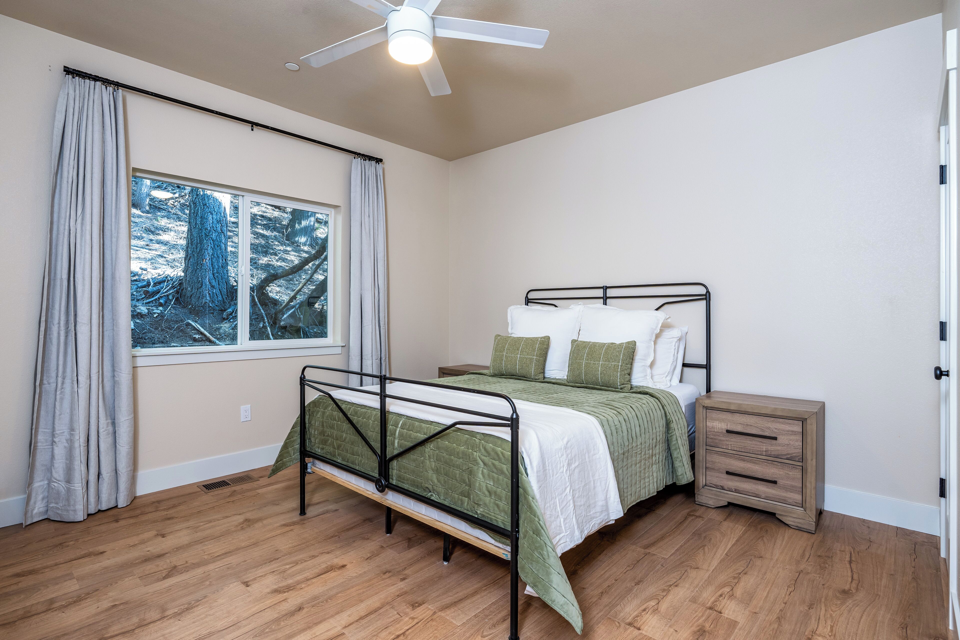 Un dormitorio con una cama, una mesita de noche, un ventilador de techo y una ventana con cortinas.