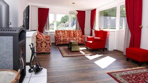 A living room with a fireplace, red and patterned sofas, a red armchair, and a large window with red curtains. - Ferienhaus Yvonne in Wendorf im Bernstein Resort (Wendorf)